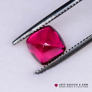 May include: A vibrant pink gemstone with a square shape, held by tweezers. The gemstone has a faceted cut, creating a sparkling effect.