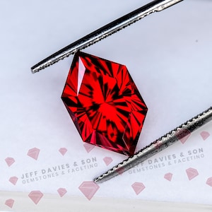 May include: A faceted, vibrant red gemstone held by metal tweezers. The gem has a complex, geometric cut, reflecting light. The background features the text "JEFF DAVIES & SON GEMSTONES & FACETING".