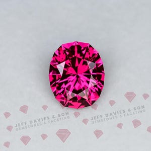 May include: A vibrant, oval-cut gemstone in a deep magenta-pink hue with intricate geometric faceting that reflects light. The stone is centred above the text JEFF DAVIES & SON GEMSTONES & FACETING, accompanied by small diamond-shaped icons.