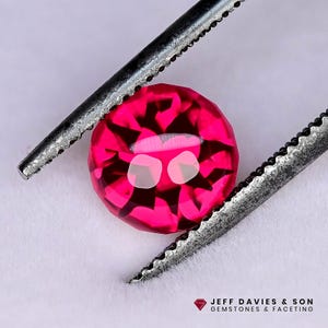 May include: A faceted, round, hot pink gemstone held by a pair of silver tweezers. The gem has a complex internal structure with light reflections. The bottom right corner features the text "JEFF DAVIES & SON GEMSTONES & FACETING".