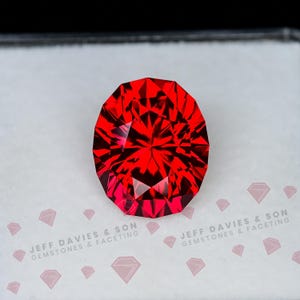 Könnte beinhalten: Ein leuchtend roter Edelstein im Ovalschliff mit komplexen Facetten, die das Licht reflektieren, präsentiert auf einer weißen Karte mit dem Text JEFF DAVIES & SON GEMSTONES & FACETING und kleinen rautenförmigen Symbolen.