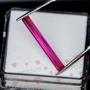 Puede incluir: Una piedra preciosa de color rosa vibrante, larga y delgada, con corte baguette, sostenida por pinzas de metal plateado. La piedra está colocada diagonalmente sobre una base de espuma blanca dentro de una caja de plástico transparente con el texto Davies & Son Gemstones & Faceting.