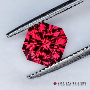 May include: A vibrant, faceted, octagonal red gemstone held by metal tweezers. The gem's intricate cuts create a starburst effect. The image is set against a white background, with the text "JEFF DAVIES & SON GEMSTONES & FACETING" at the bottom.