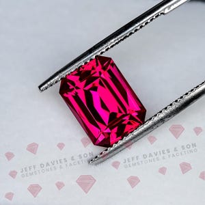 May include: A vibrant, emerald-cut gemstone with a deep, saturated pinkish-red hue held by silver-toned metal tweezers. The stone features precise, multi-faceted surfaces that reflect light, creating sharp geometric patterns across its surface.