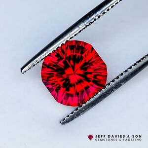 Puede incluir: Una gema facetada, octogonal, de color rojo vibrante, sujeta con unas pinzas plateadas. El interior de la gema presenta un patrón oscuro e intrincado. La imagen incluye el texto "JEFF DAVIES & SON GEMSTONES & FACETING".