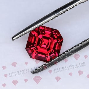 May include: A vibrant red, faceted gemstone with a geometric, multi-layered square cut held by silver-toned metal tweezers. The stone displays deep crimson hues and sharp, symmetrical edges. The text JEFF DAVIES & SON GEMSTONES & FACETING is visible.