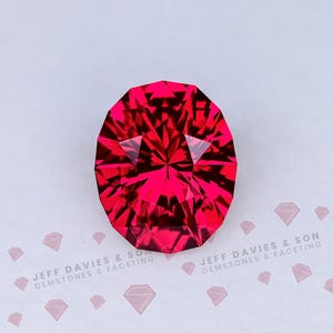 May include: A faceted, oval-shaped gemstone with a vibrant red hue. The gem's surface reflects light, creating a starburst effect. The gemstone is set against a white background with the text "JEFF DAVIES & SON GEMSTONES & FACETING".