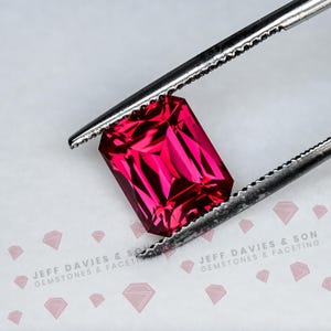 Könnte beinhalten: Ein leuchtender, rechteckig geschliffener Edelstein mit einer tiefen, gesättigten magentaroten Farbe und facettierter Oberfläche, gehalten von einer Metallpinzette. Der Text JEFF DAVIES & SON GEMSTONES & FACETING ist unten zu sehen.