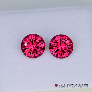May include: Two round, faceted, red gemstones on a white surface. The gemstones are approximately 6mm in diameter.