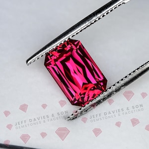 May include: A vibrant, rectangular-cut, deep pink gemstone held by silver-toned metal tweezers. The stone features a faceted surface that reflects light, creating sharp geometric patterns. The text JEFF DAVIES & SON GEMSTONES & FACETING is visible.