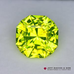 May include: A faceted, square-cut gemstone with a vibrant, lime green hue. The gem's multiple facets reflect light, creating a brilliant, sparkling effect. The gemstone is set against a neutral background, highlighting its color and clarity. The logo "JEFF DAVIES & SON GEMSTONES & FACETING" is in the bottom right corner.