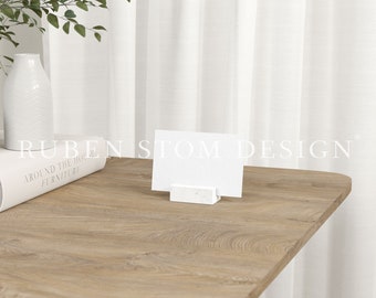 May include: A white card with the text "RUBEN STOM DESIGN" is displayed on a white marble card holder. The card holder is sitting on a light brown wooden table. A white book with the text "AROUND THE HOME FURNITURE" is on the table.