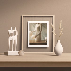 May include: A framed photograph of a person in a white dress holding a glass orb. The photo is on a wooden table with a white sculpture and a vase with dried flowers.