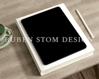 May include: A white tablet computer with a black screen is lying on a white book. A white stylus pen is lying on the book to the right of the tablet. The book and tablet are on a light brown wooden surface.