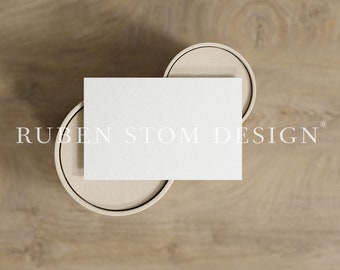 May include: A white blank card mockup on a light brown wooden surface. The card is sitting on top of two beige round objects.