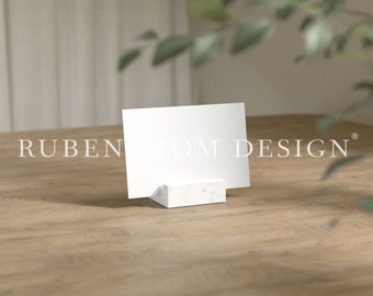 May include: A white card mockup on a white marble stand, sitting on a wooden table. The card is blank and ready for your design.