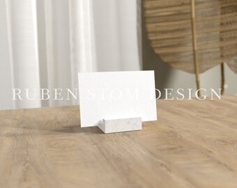 May include: A white card holder with a blank white card on top. The card holder is made of marble and has a textured surface. The card holder is sitting on a wooden table.