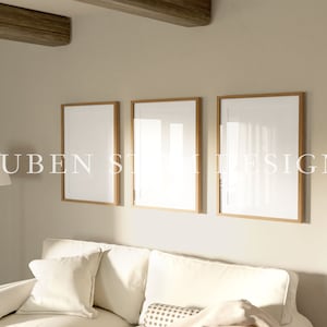 May include: Three empty picture frames with a light wood frame are hung on a beige wall above a white sofa. The frames are arranged in a row, with the leftmost frame slightly higher than the other two. The sofa is covered in a white fabric and has two white cushions. The wall is painted a light beige colour. The room is lit by a floor lamp with a white shade.