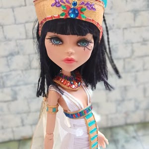Puede incluir: Muñeca vestida como faraón egipcio, con una corona dorada con detalles turquesas y rojos. La muñeca tiene pelo negro, ojos azules y un vestido blanco con adornos coloridos. La muñeca mide aproximadamente 30 cm.