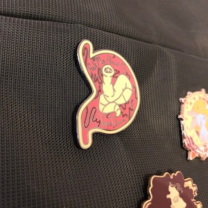 May include: A gold-colored enamel pin with a red stomach-shaped outline. Inside the outline is a yellow worm with a face and the text "Ask Me About My Tapeworm".