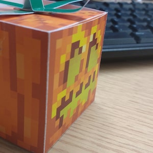 May include: A small orange and yellow cardboard box shaped like a Minecraft pumpkin with a green ribbon.