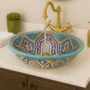 Puede incluir: Un lavabo de baño decorativo con un vibrante patrón azul, amarillo y morado. El lavabo está sobre una encimera blanca con un grifo dorado, espejo y dispensador de jabón. También se ven una pequeña planta verde y una pila de toallas blancas.