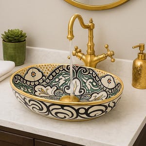May include: A decorative bathroom sink with a gold faucet, a round gold mirror, and a gold soap dispenser. The sink has a black, white, and green floral pattern. A small green plant and white towels are also visible.