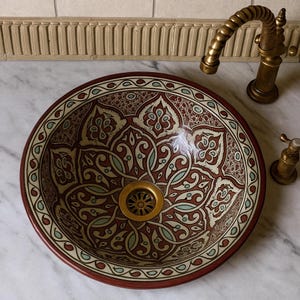 Op de afbeelding: Ronde keramische wastafel met ingewikkelde, symmetrische patronen in bordeauxrood, crème en teal. De wastafel heeft een messing afvoer en staat op een marmeren aanrecht. Een bijpassende messing kraan en handvat zijn zichtbaar.