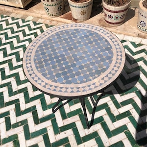 Puede incluir: Una mesa redonda con una tapa de mosaico azul y blanco y una base de metal negro. La mesa está sobre un suelo de baldosas verdes y blancas.