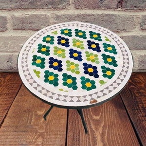Puede incluir: Una mesa redonda de mosaico con fondo blanco y un patrón de flores verdes, azules y amarillas. La mesa está hecha de azulejos pequeños y tiene un borde blanco.