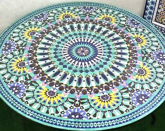 Op de afbeelding: Een ronde tafelblad met een kleurrijk mozaïekontwerp. Het ontwerp heeft een centraal stervormig patroon omgeven door herhalende geometrische vormen in tinten blauw, groen, geel en wit.