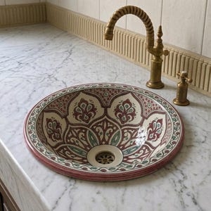 Pode incluir: Uma pia redonda de cerâmica decorativa com um intrincado padrão floral em vermelho, creme e verde. A pia está embutida em uma bancada de mármore, com uma torneira de latão e acessórios correspondentes. O design da pia lembra a arte marroquina tradicional.