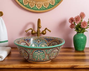 May include: A decorative ceramic sink with a detailed floral pattern in blue, green, and brown. The sink is set on a wooden surface with a brass faucet. A green vase with pink flowers is to the right.