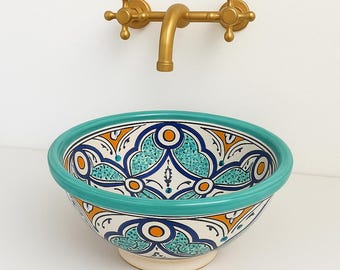 May include: A decorative ceramic sink with a teal rim and intricate blue, orange, and white patterns. Above, a gold-colored faucet with cross-shaped handles is mounted on the wall. The sink is round and sits on a small, off-white base.