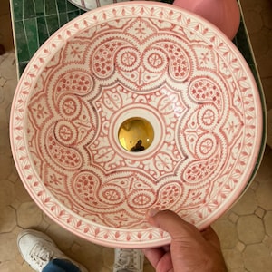 May include: A round, decorative ceramic sink with a detailed, symmetrical pattern in shades of red and white. The sink has a gold-colored drain and a light pink rim. The design is reminiscent of traditional Moroccan or Middle Eastern art.