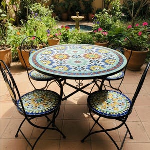 May include: A round outdoor dining set with a mosaic tile tabletop and matching chair seats. The table and chairs have black metal frames. The mosaic design features blue, yellow, and green floral patterns. The set is surrounded by potted plants.