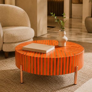 Könnte beinhalten: Ein runder Couchtisch mit einem Muster aus abwechselnden vertikalen orangefarbenen und naturfarbenen Holzstreifen. Der Tisch steht auf drei zylindrischen Metallbeinen und trägt ein rechteckiges beiges Buch sowie eine kleine weiße Keramikvase mit Grünpflanzen.