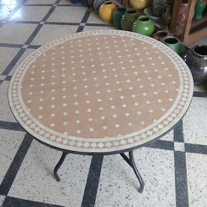 May include: A round, terracotta-colored table with a white geometric tile pattern and a black metal frame. The table is sitting on a black and white checkered floor.