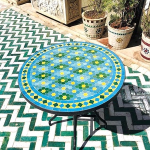 Puede incluir: Una mesa redonda de mosaico con un patrón geométrico azul, verde y amarillo. La mesa tiene un marco de metal negro y tres patas. La mesa está sobre un suelo de baldosas blancas y verdes.