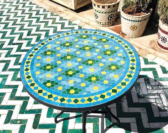 Op de afbeelding: Een ronde mozaïektafel met een geometrisch patroon in blauw, groen en geel. De tafel heeft een zwart metalen frame en drie poten. De tafel staat op een witte en groene betegelde vloer.