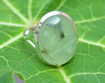 May include: A silver ring with a large, oval, light green gemstone. The gemstone has inclusions and is set in a polished silver band. The ring is displayed on a green leaf.