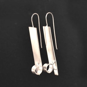 May include: A pair of silver earrings with a long, rectangular shape and a curved, circular detail at the bottom.