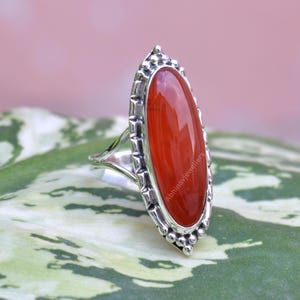 May include: A silver ring with an oval, polished, orange-red gemstone. The ring has a detailed, textured silver band and a decorative bezel setting around the stone. The ring is set against a green and pink background.