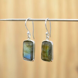Puede incluir: Un par de pendientes de plata con piedras rectangulares de labradorita. Las piedras tienen un brillo iridiscente verde, azul y dorado.