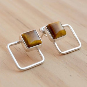 以下が含まれることがあります： 正方形のフレームと茶色のタイガーアイストーンのアクセントが付いたシルバーのイヤリングのペアです。