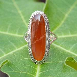 May include: A silver ring featuring an oval, polished, reddish-brown gemstone. The gemstone is set in a detailed, textured silver band. The ring is displayed on a green leaf, showcasing the jewelry's design and color.