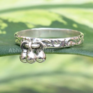 Puede incluir: Un anillo de plata con un diseño floral y tres pequeñas campanas colgantes. El anillo está sobre un fondo verde y blanco. El texto "Abhi Jewellers" es visible en el anillo.