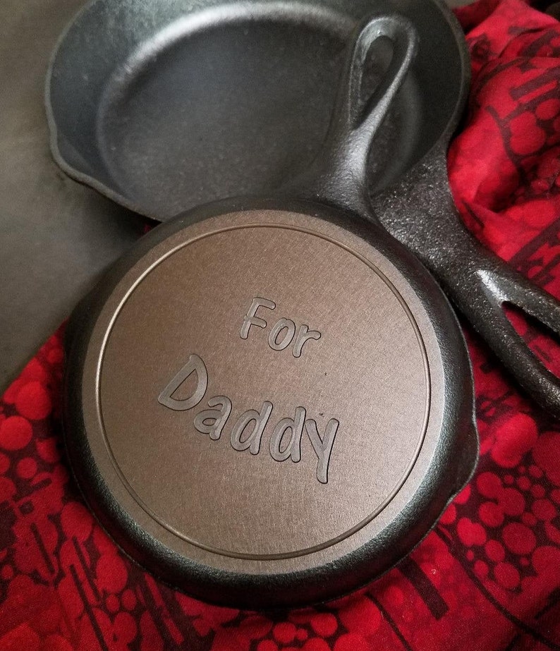 Może przedstawiać: Dwie czarne żeliwne patelnie. Mniejsza patelnia ma wygrawerowane na spodzie słowa "For Daddy".