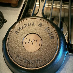 Peut inclure: Une poêle en fonte noire avec une poignée, personnalisée avec les noms "Amanda & John" et la date "6♡13♡15".