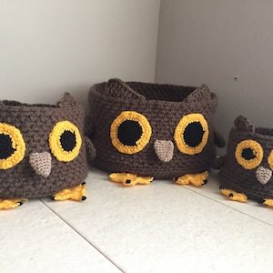May include: Three crocheted owl-shaped baskets in different sizes. The largest basket is brown with yellow eyes and a brown beak. The medium basket is brown with yellow eyes and a brown beak. The smallest basket is brown with yellow eyes and a brown beak.